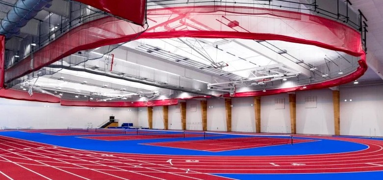 A spacious indoor track and field facility with multiple lanes of a red running track and blue field sections. Overhead, a red netting barrier hangs from the ceiling. The area is well-lit with bright overhead lights and minimalistic wooden and white walls.